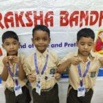 Three young schoolboys in beige uniforms with blue lanyards pose in front of a banner at a school event holding small circular badges and prize slips